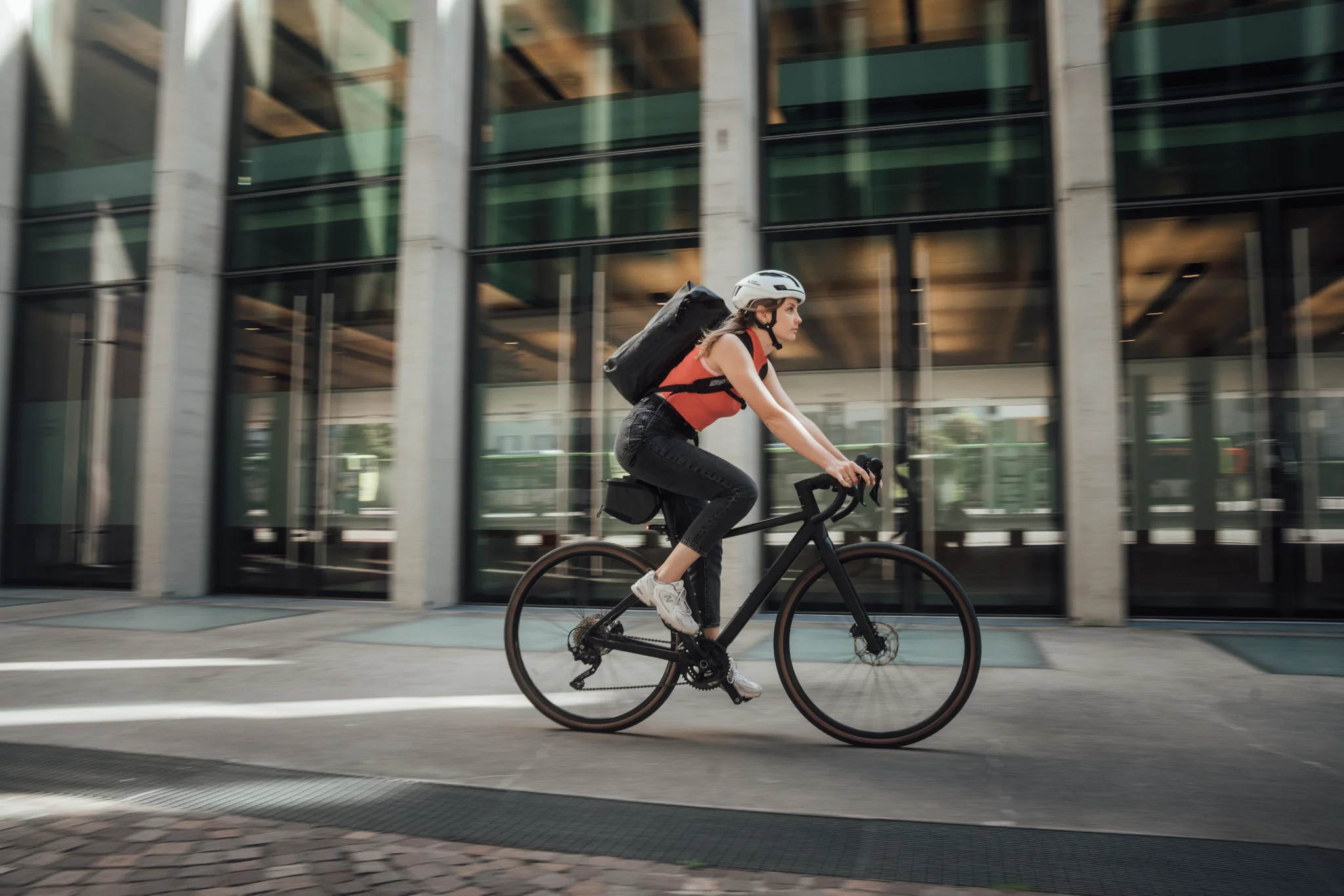 A cyclist with a black Tatonka roll-top backpack rides past a glass facade in the city