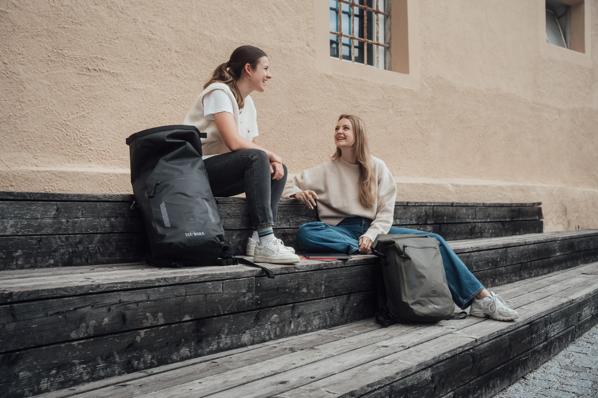 Two young women sit on wooden steps with a black and an olive Tatonka roll-top backpack