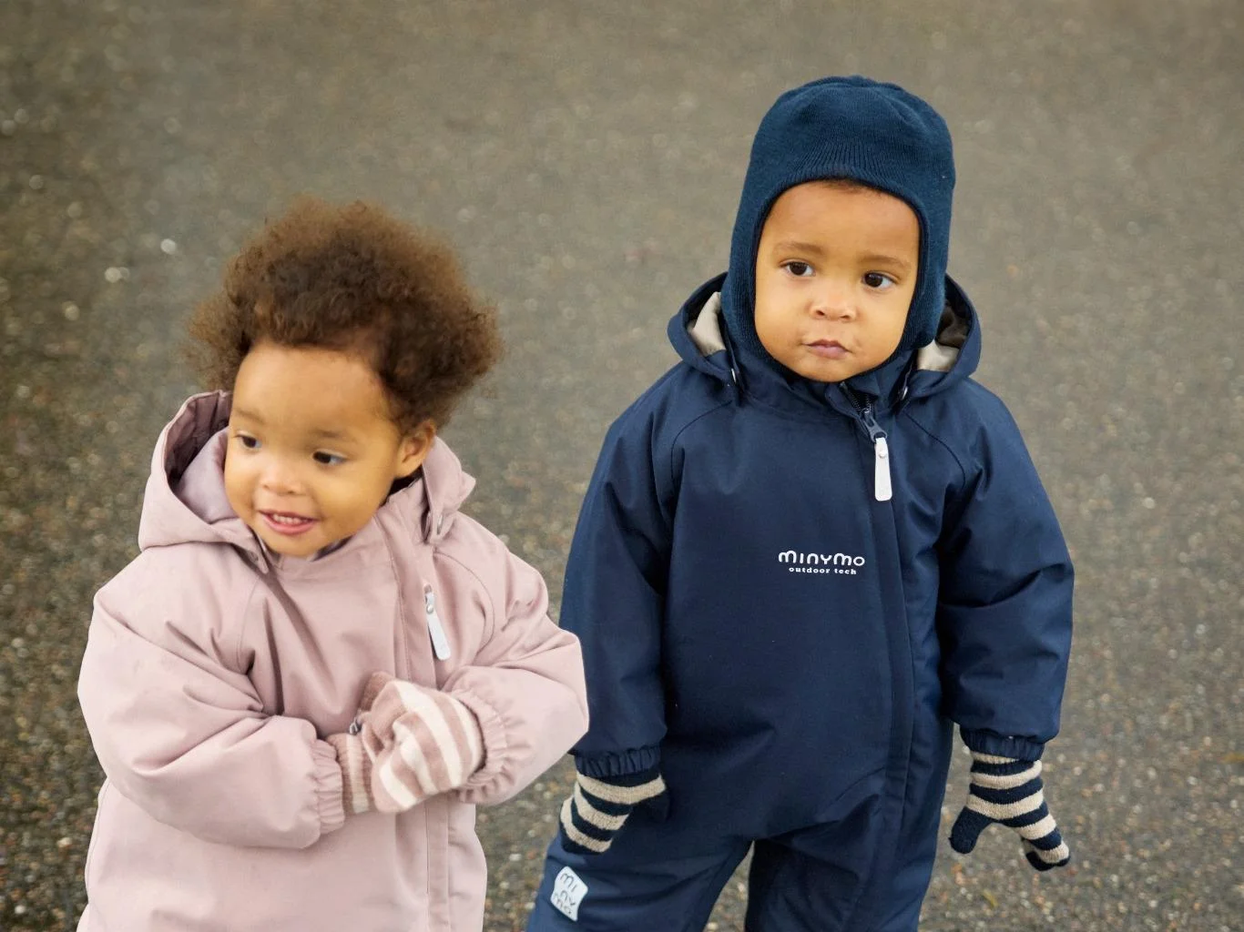 Two toddlers in waterproof Minymo outdoor suits and knitted hats stand outside on a wet autumn day