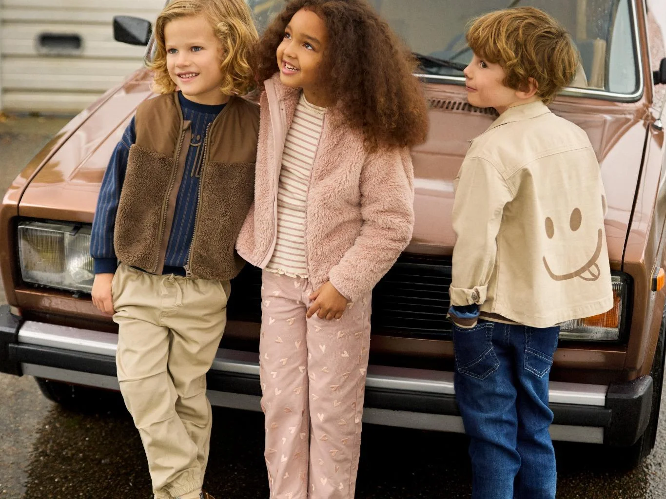 Three children in Minymo jackets and trousers stand together in front of a car on a damp autumn day