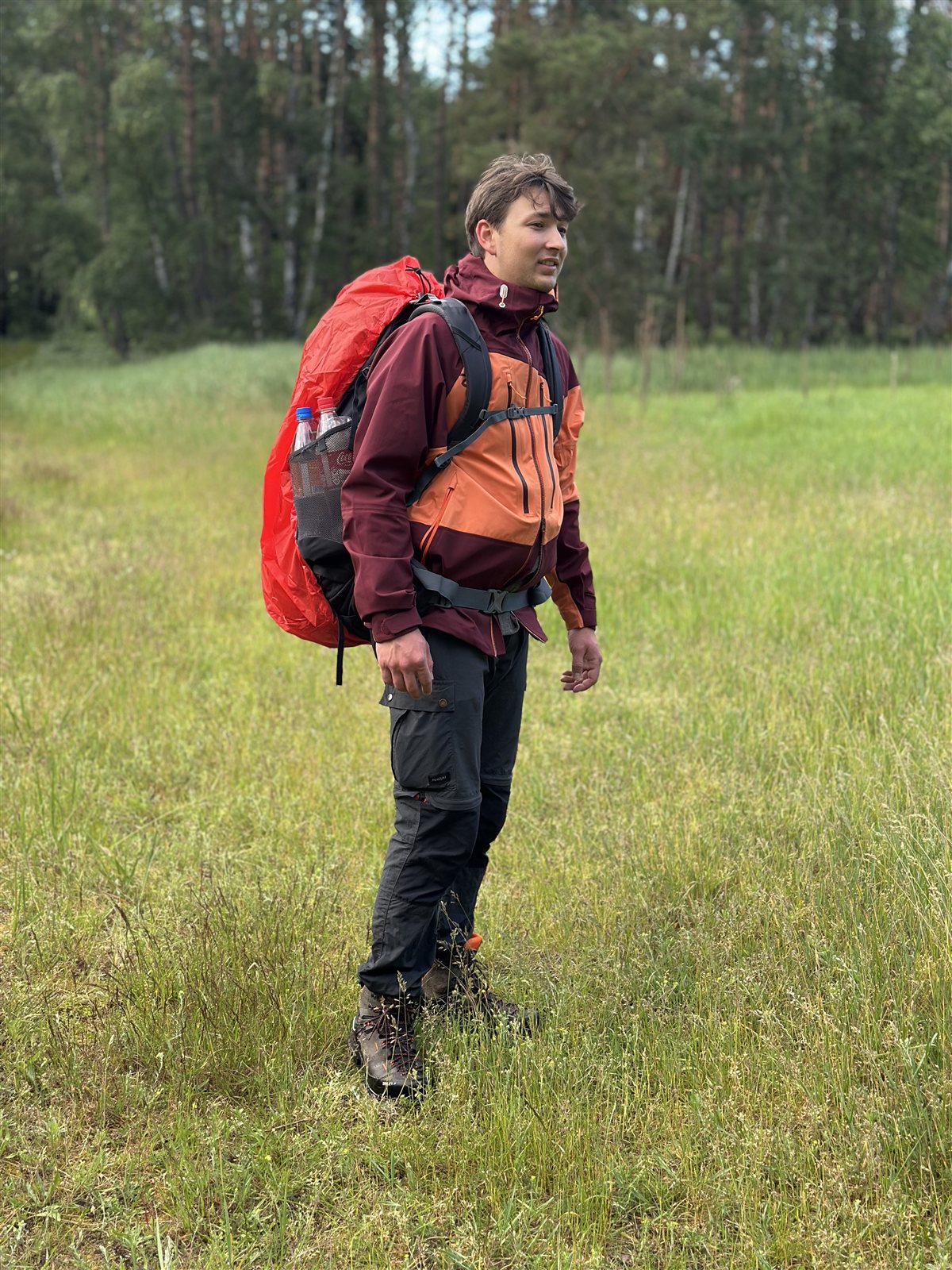 Product tester with a rucksack in the Ternua Klimmer Jacket on a meadow at the edge of a forest